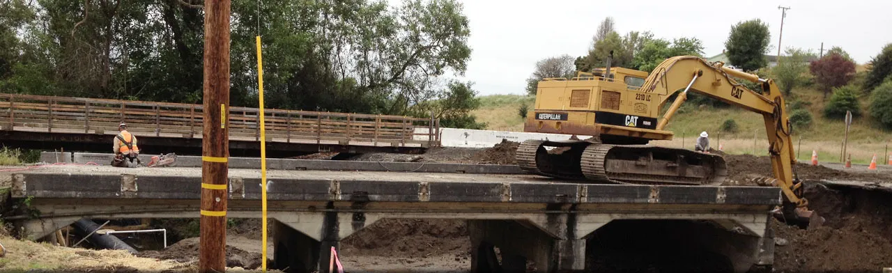 An excavator working from a bridge deck under construction alongside an existing timber bridge