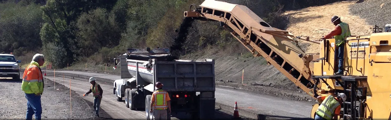 A cold planer discharging milled asphalt into a dump truck while the crew works the shoulder